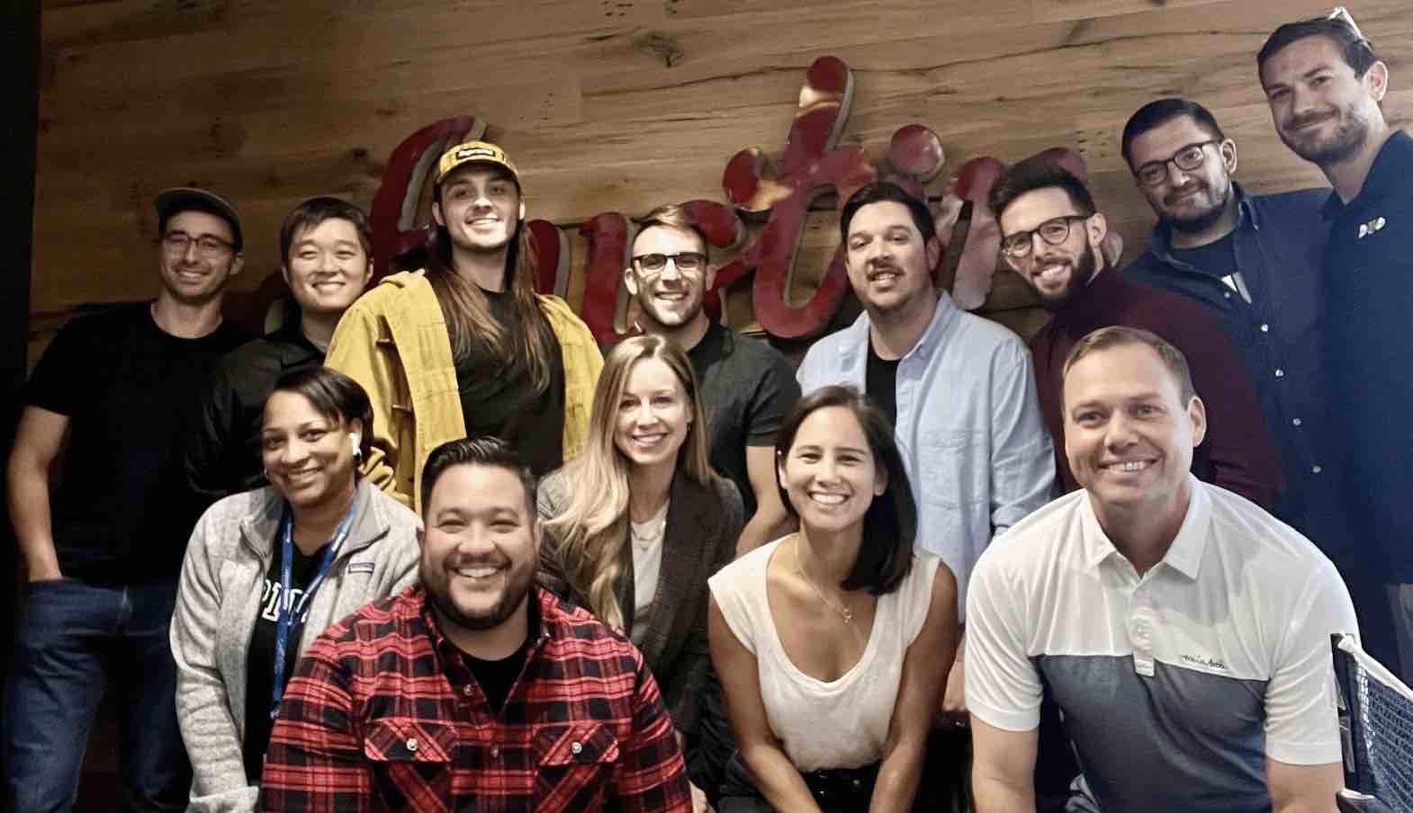 Esther Kho poses with a 12-person team in front of a wooden wall featuring a sign that reads 